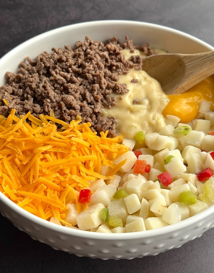 Frozen potatoes, browned ground beef, canned soup, and shredded cheddar cheese in a bowl ready to be mixed. 