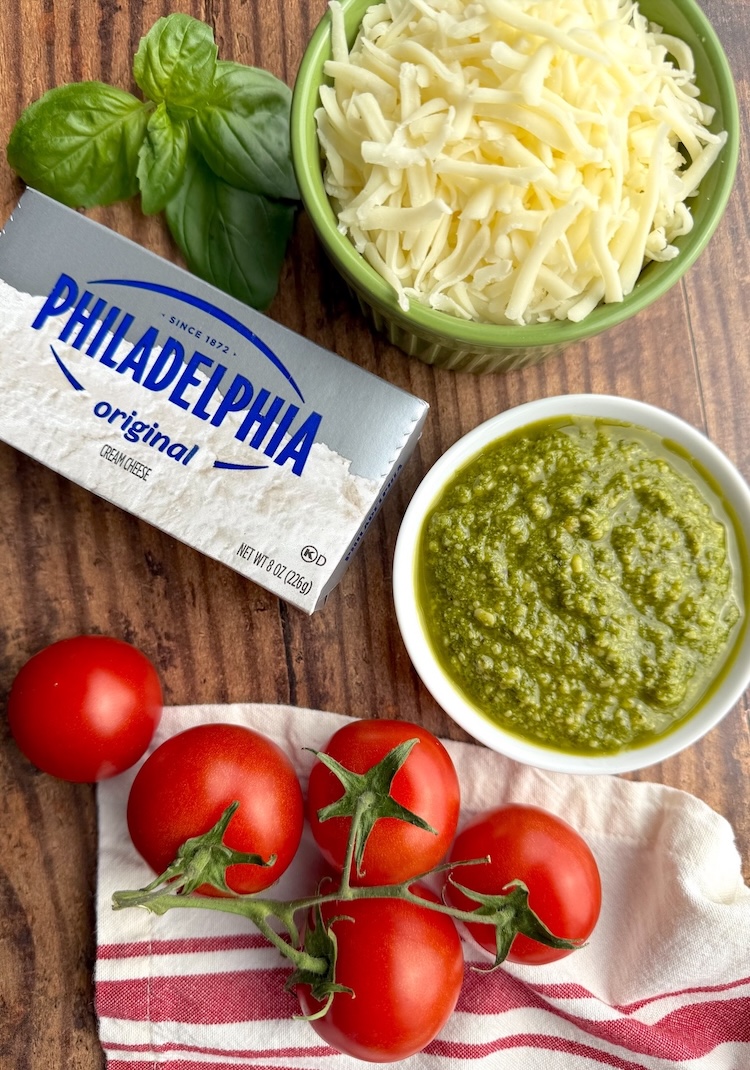 List of ingredients need to make creamy pesto baked chicken including chicken breasts, cream cheese, basil pesto, mozzarella, fresh basil, and tomatoes. 