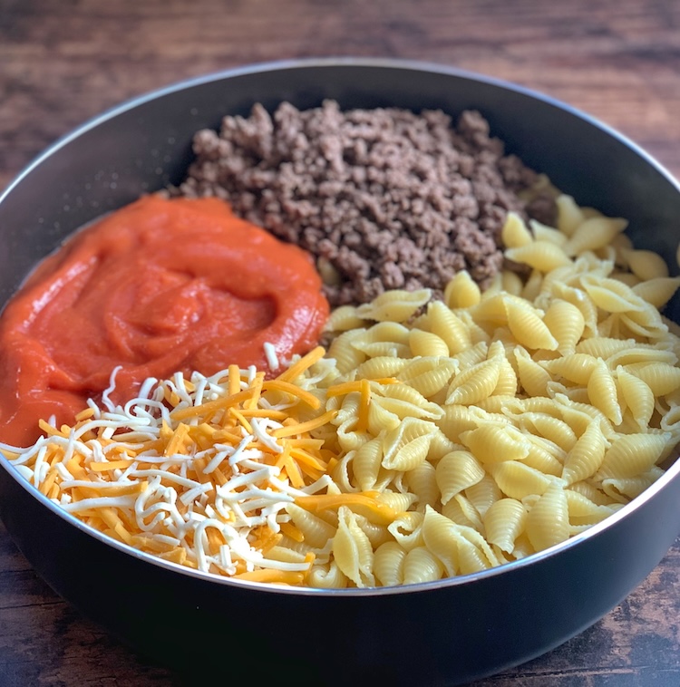 Pasta shells, ground beef, shredded cheese, and tomato soup in a pan ready to be mixed and placed into a large casserole dish for dinner. 