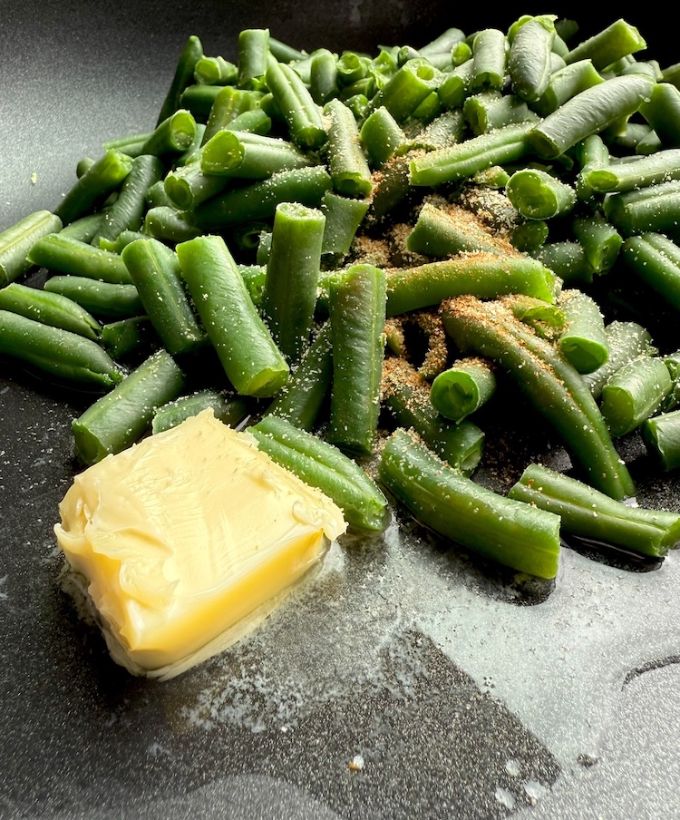 Steamed frozen green beans in a cooking pan with oil, butter, and seasoning getting ready to sauté. 